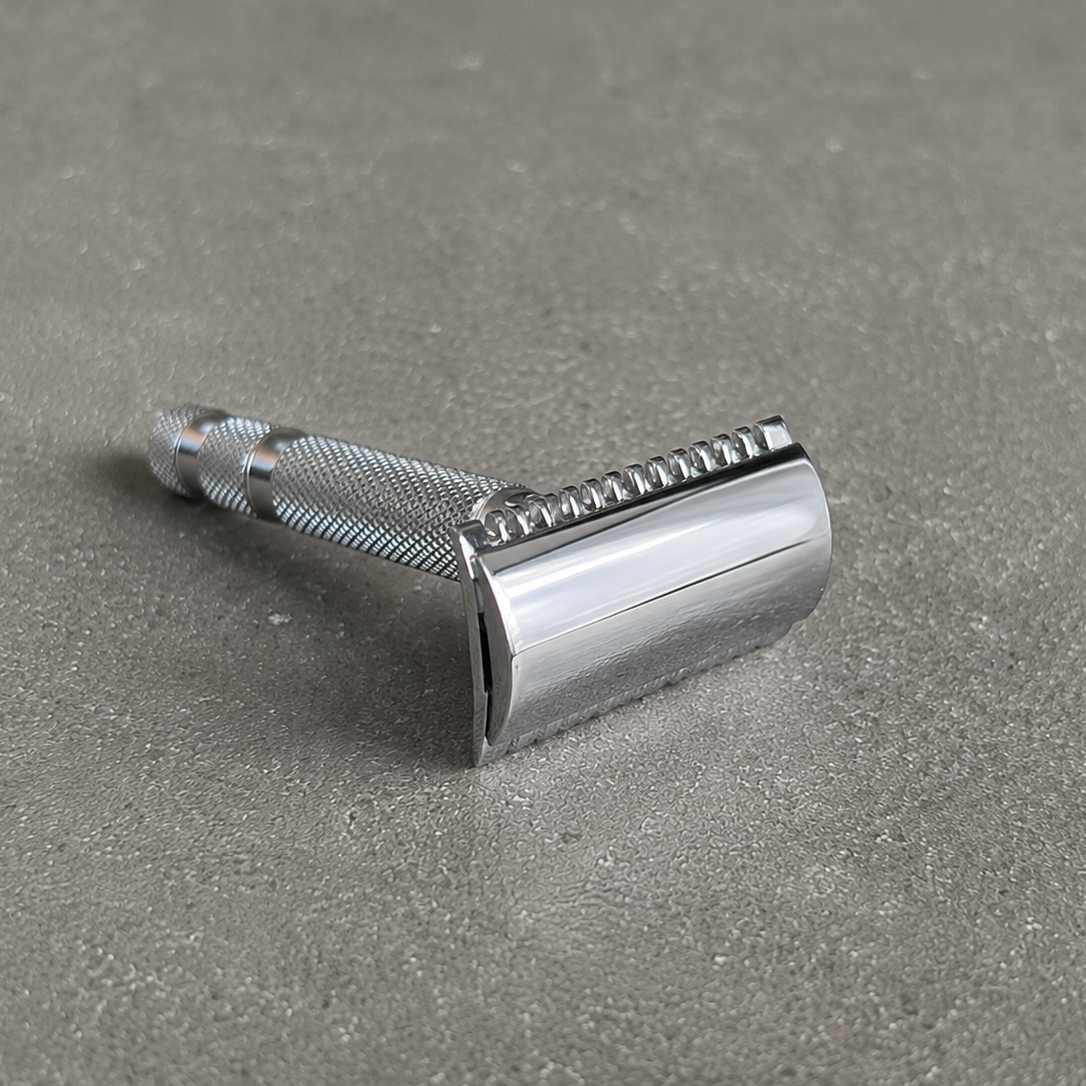 A classic stainless steel safety razor with a textured grip and polished chrome blade cover, resting on a textured gray surface.