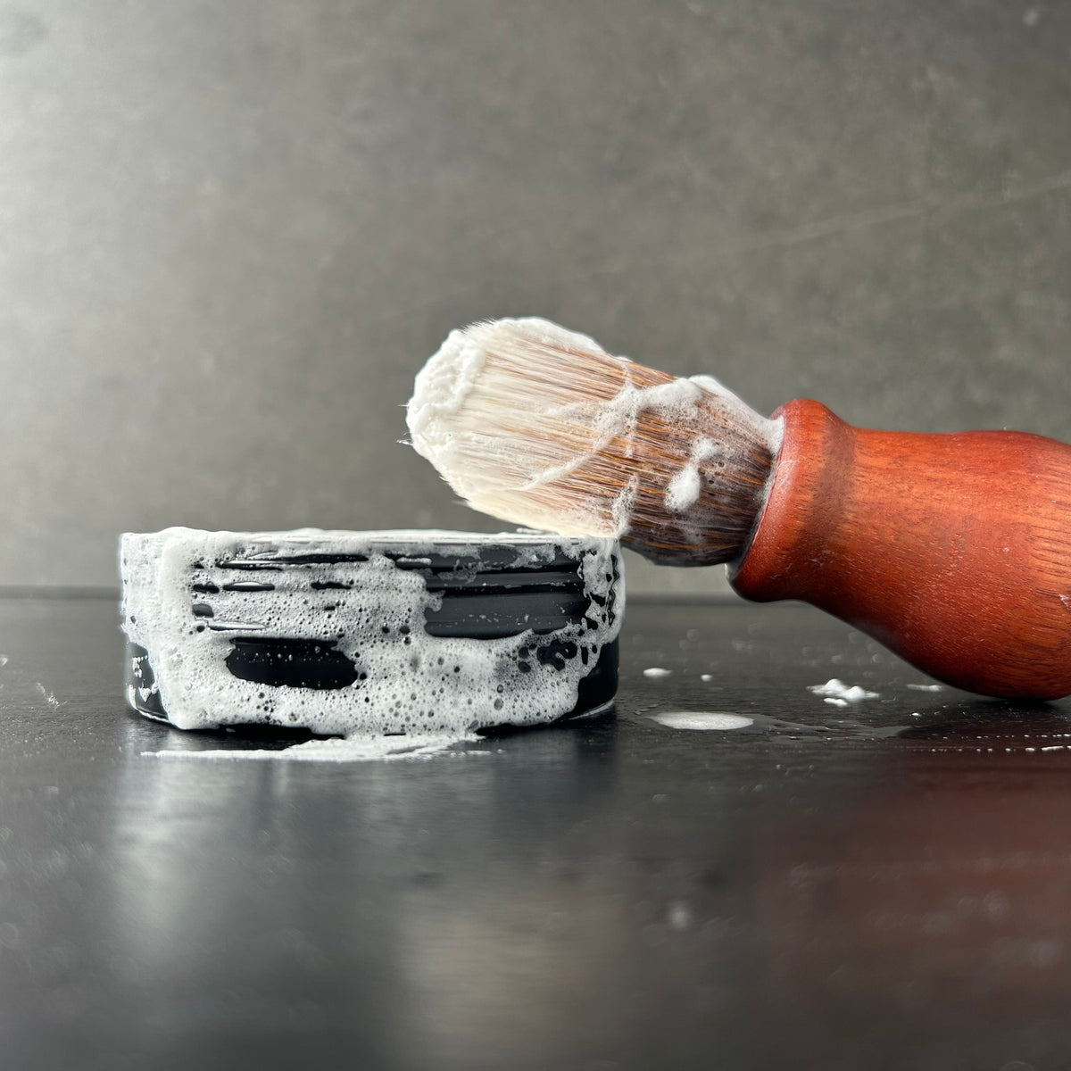 A close-up of a shaving brush with a reddish-brown wooden handle and white bristles, covered with soap lather, positioned against a dark gray surface next to a black rectangular object with white soap lather.